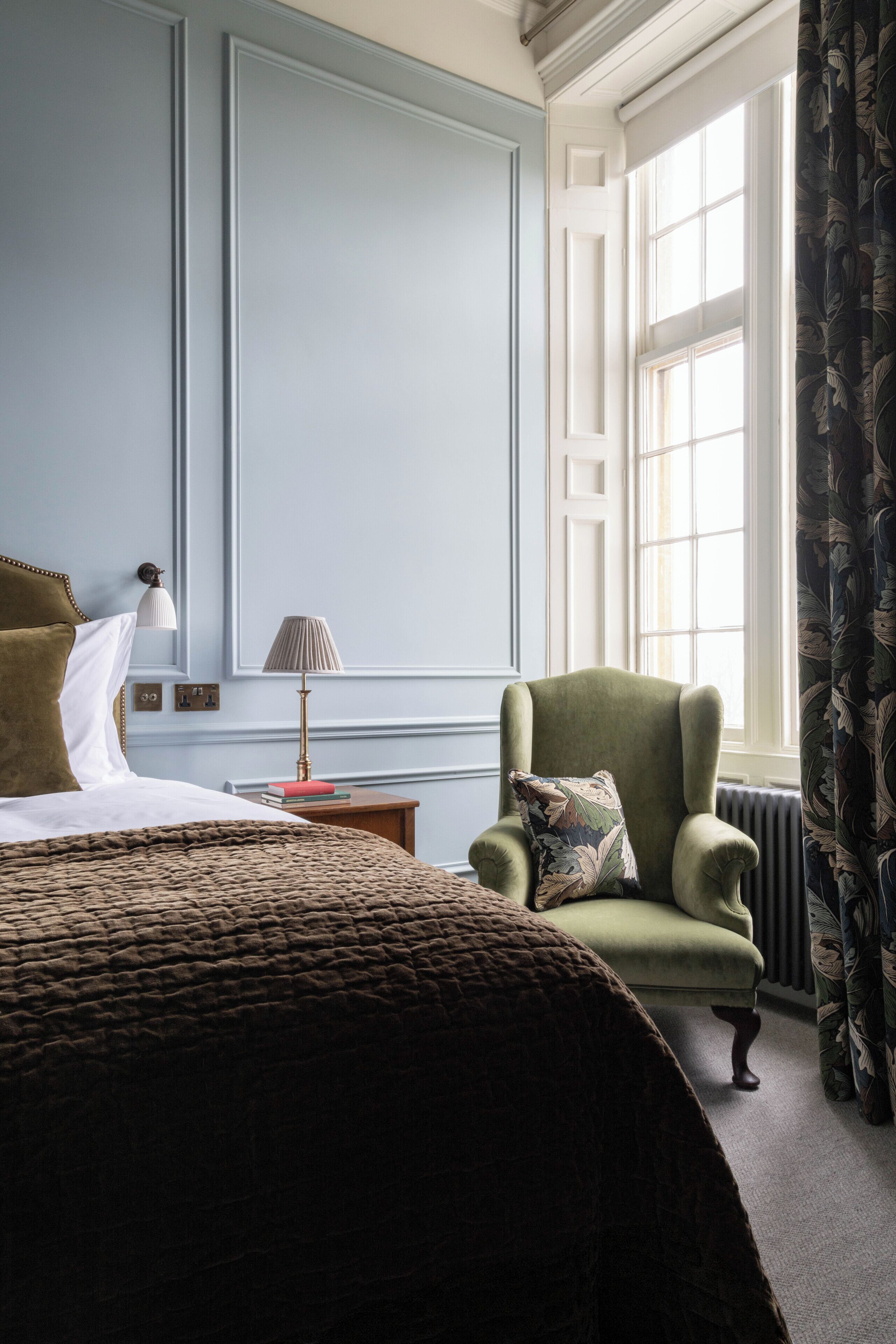 A well-lit room with a bed, a green armchair, a nightstand with a lamp, and a window with curtains.