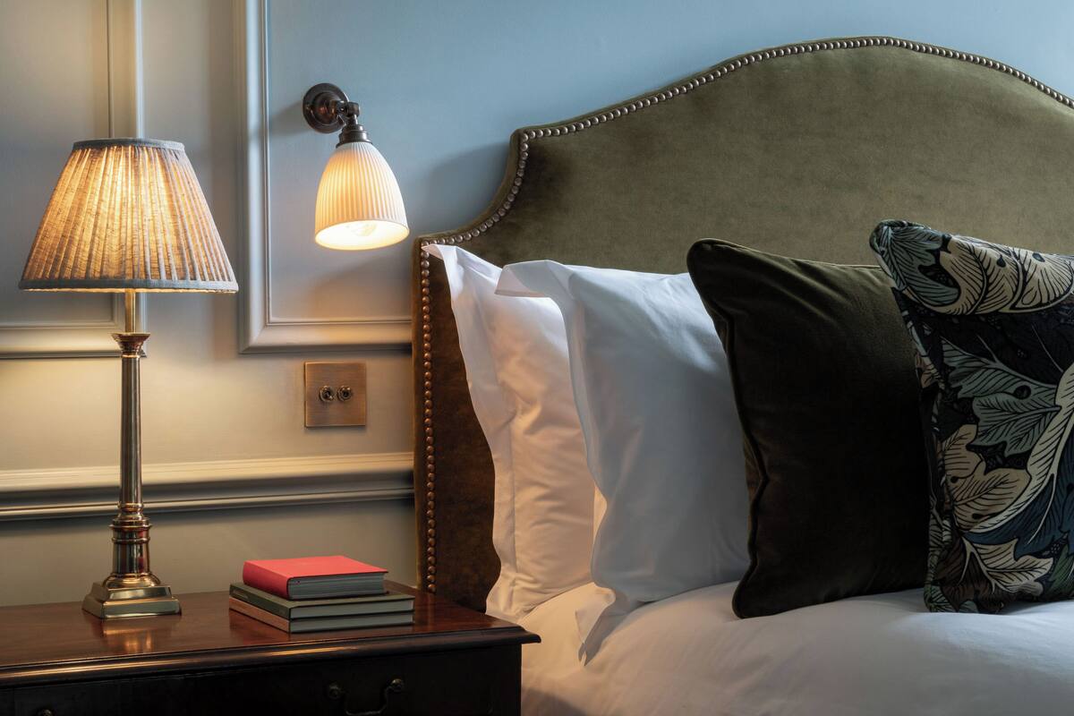 A neatly made bed with a headboard, a bedside lamp, and a nightstand with books.
