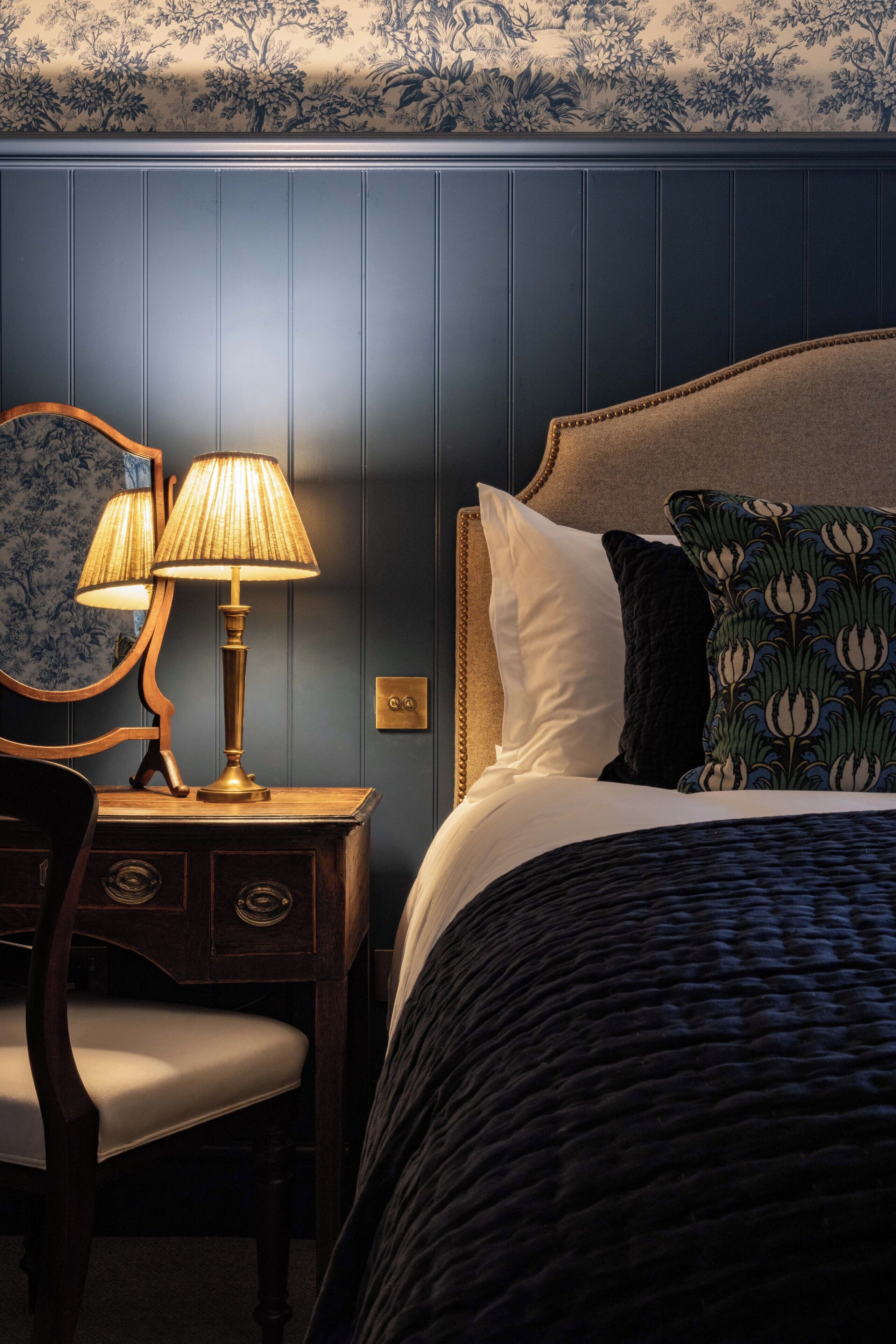 Una cama bien hecha con un edredón oscuro, una mesita de noche de madera con una lámpara y una silla.