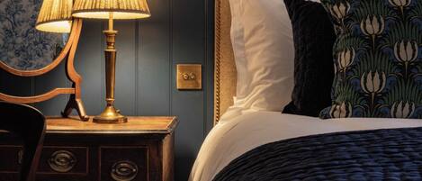 Una cama bien hecha con un edredón oscuro, una mesita de noche de madera con una lámpara y una silla.