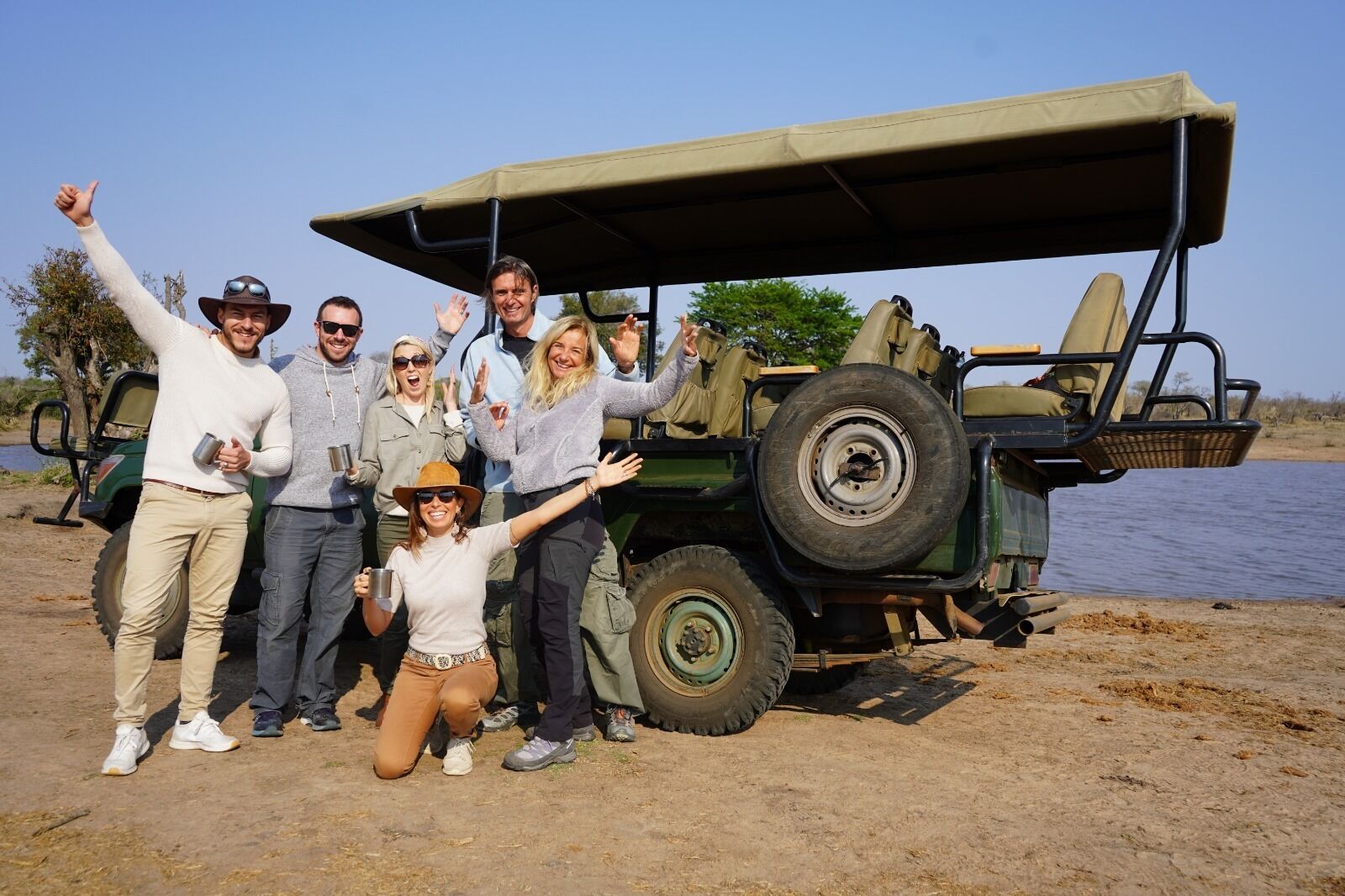 A group of people posing in front of a safari vehicle with a canopy, near a body of water.