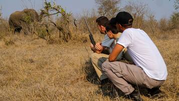 A group of people in a natural setting with dry grass and trees, observing an elephant in the distance.