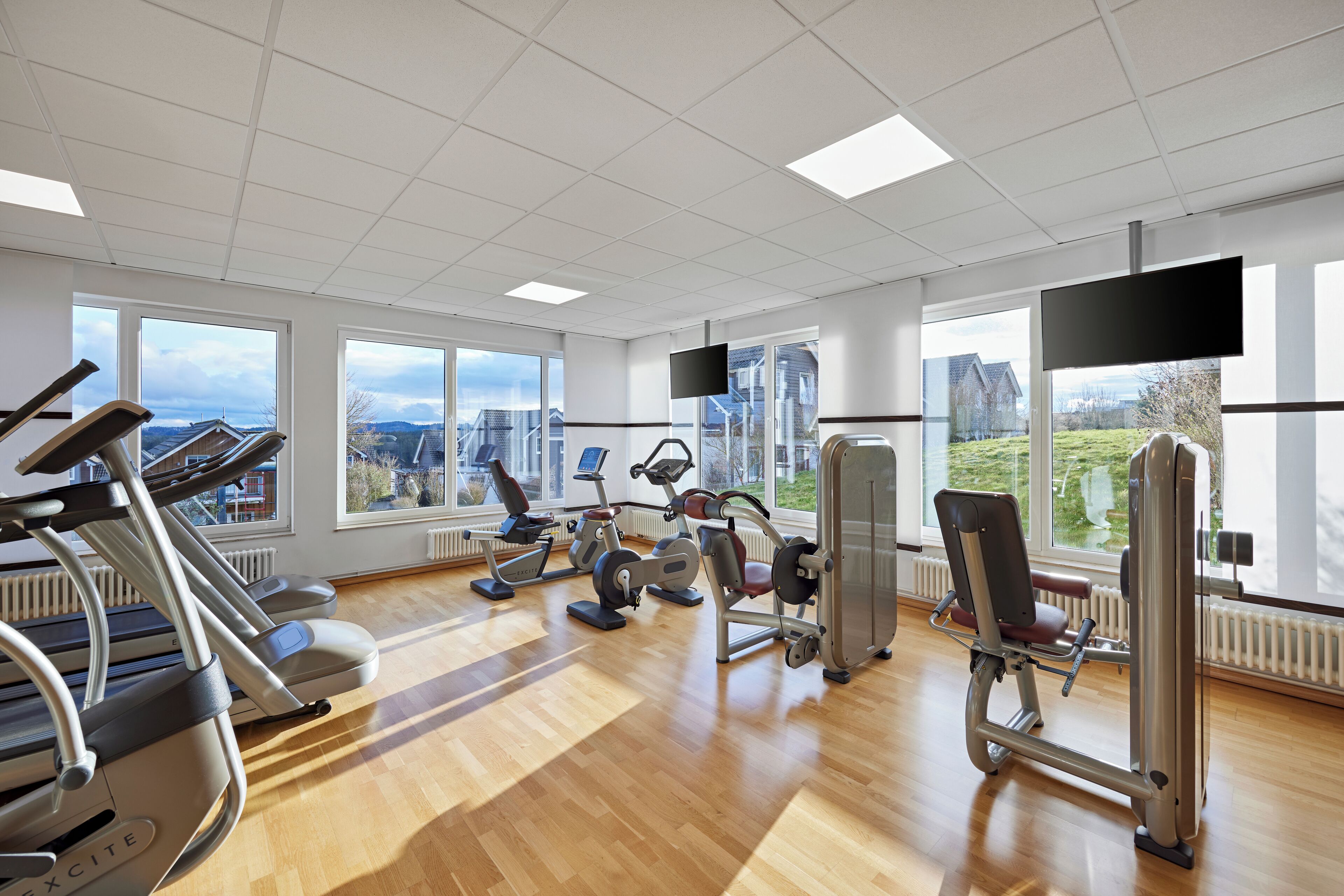 A gym with cardio machines, weight training equipment, and large windows offering a view of the outdoors.
