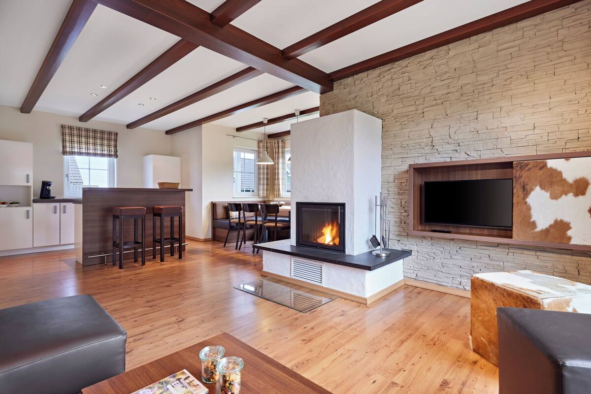 A modern living room with a fireplace, wooden beams, stone wall, and a TV mounted on the wall.