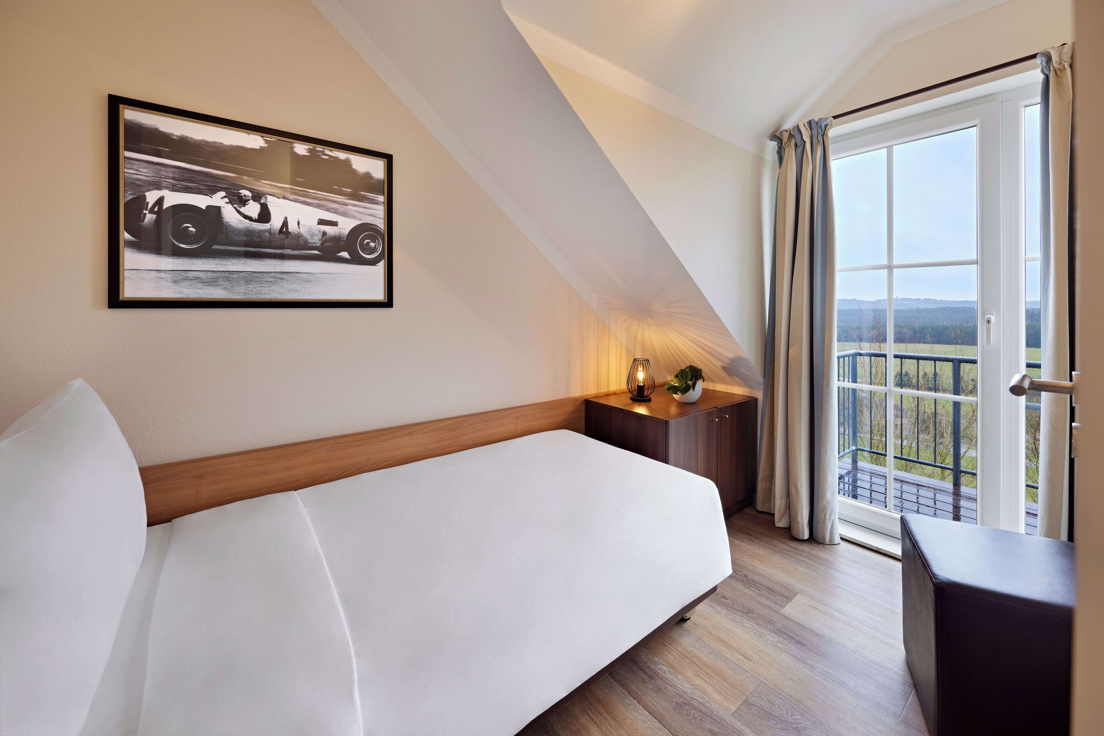 A modern hotel room with a large bed, a side table with a lamp, a framed picture on the wall, and a balcony with a view.