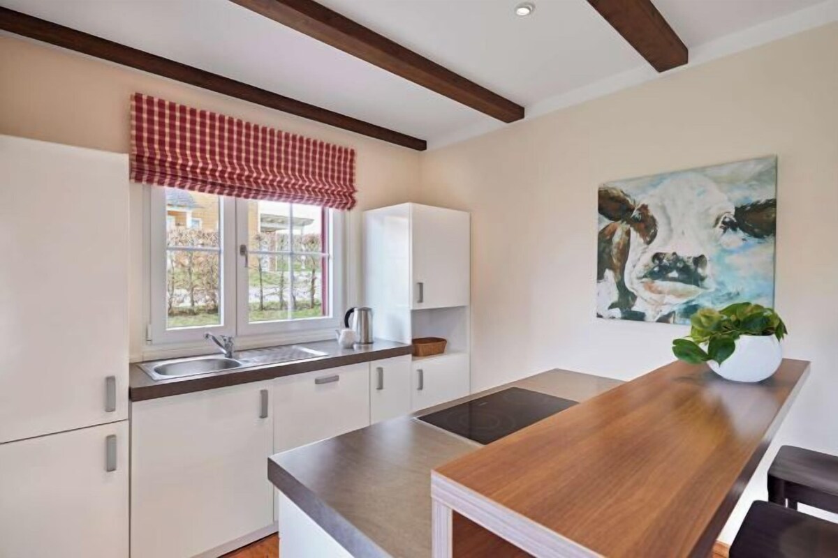 A kitchen with white cabinets, a wooden countertop, a window with a red valance, and a painting on the wall.