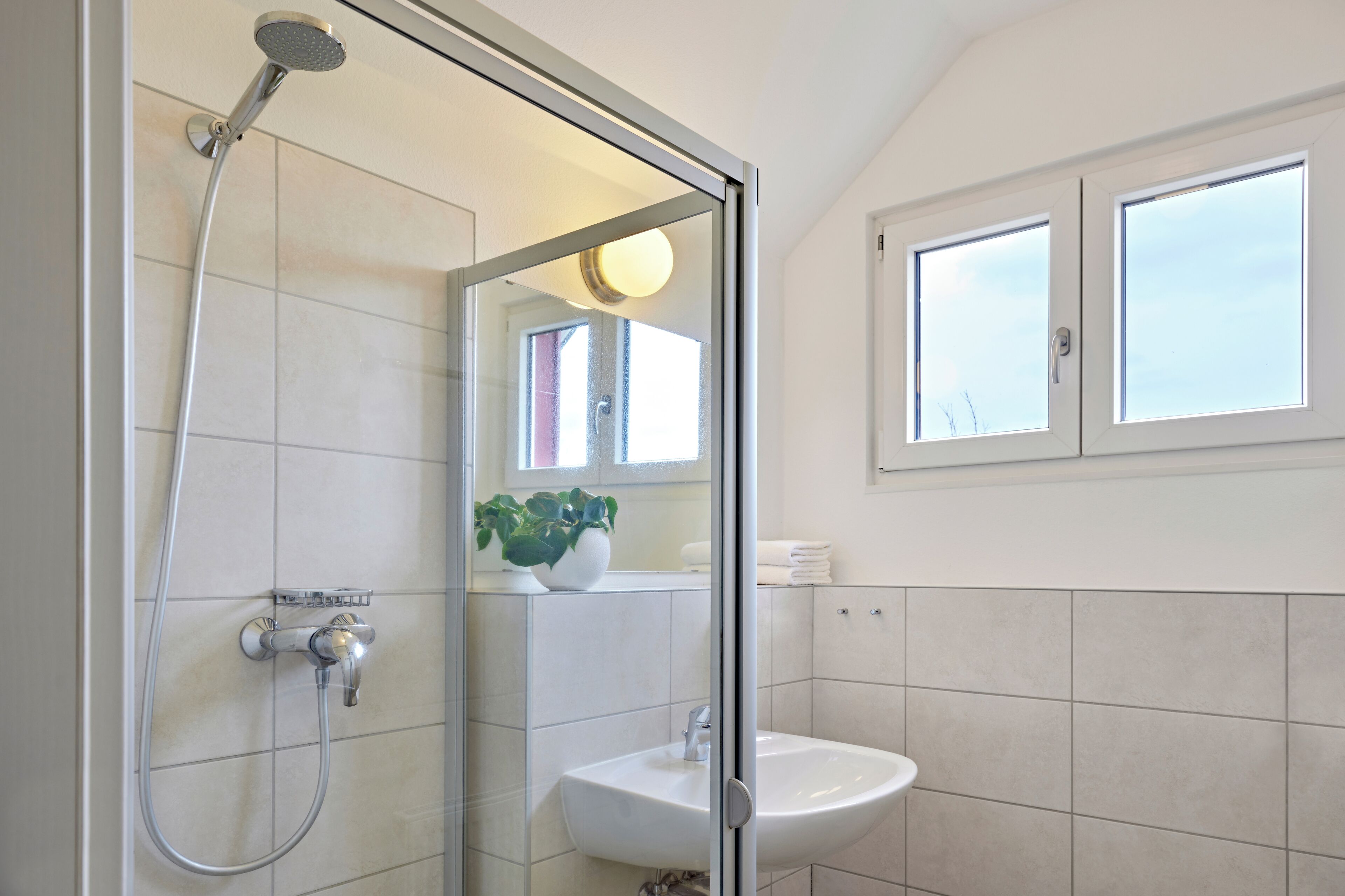 Un baño moderno con ducha envuelta en vidrio, un lavamanos blanco y una ventana sobre el espejo.