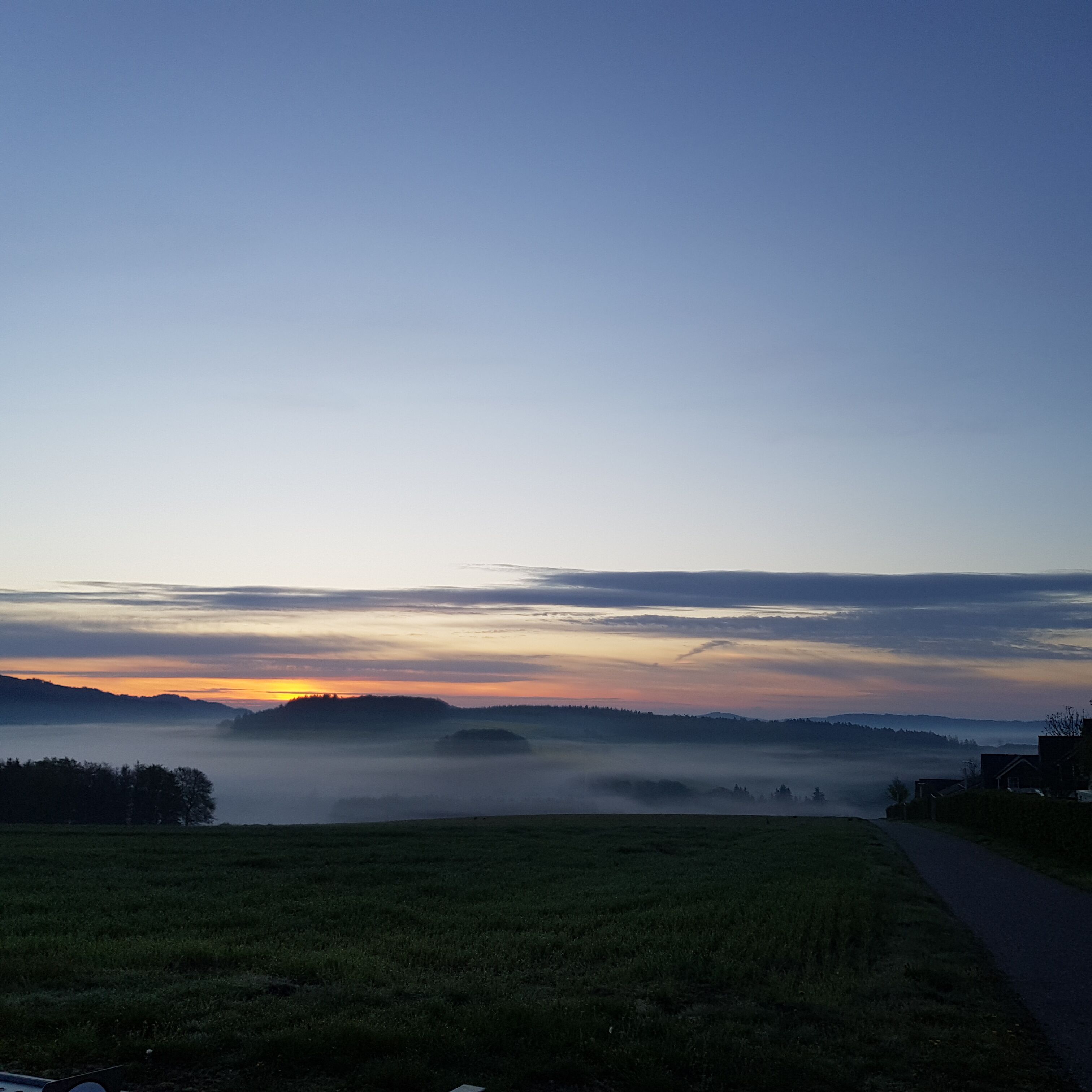 A rural landscape with a road, fields, and a sunrise or sunset over hills.