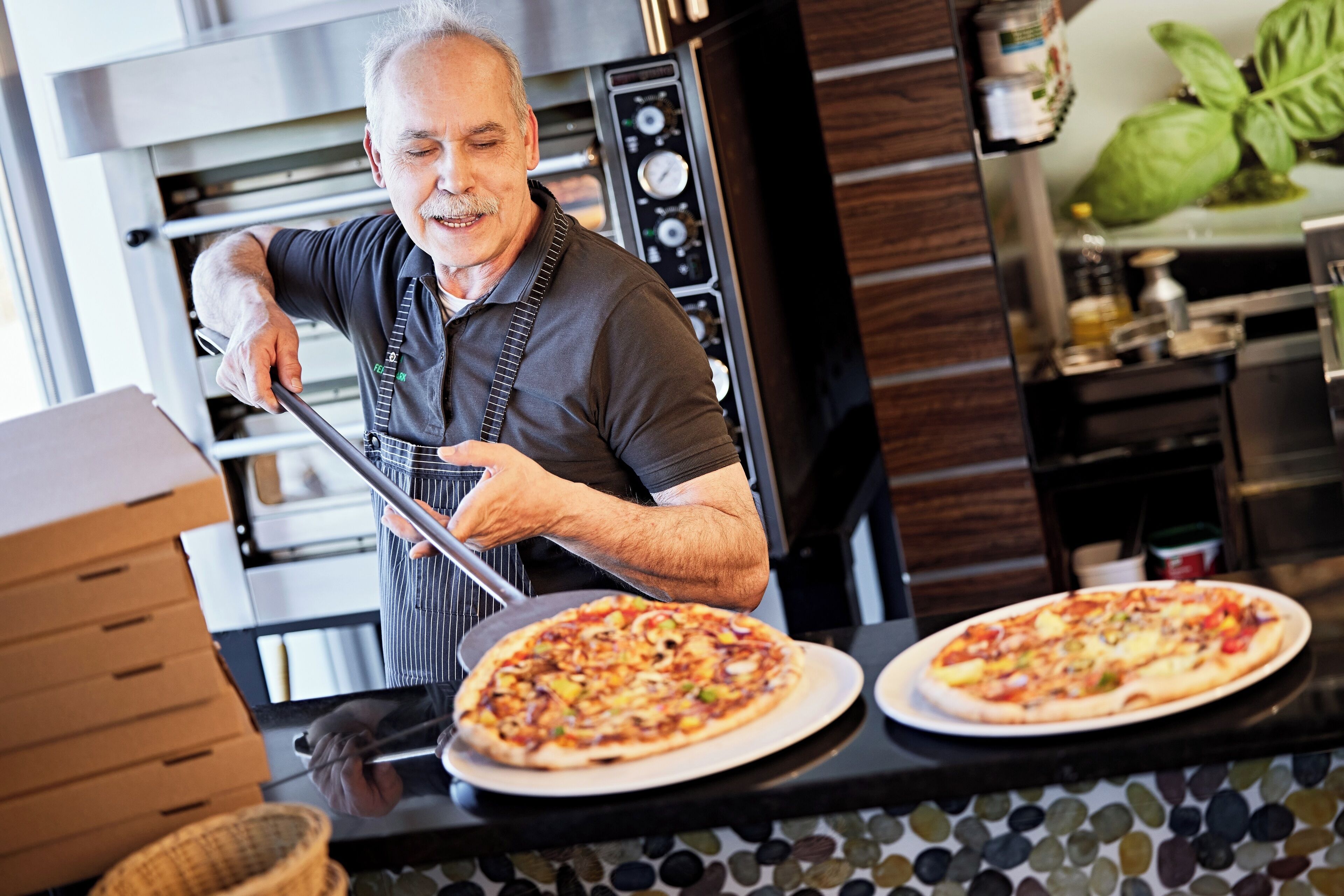 Une cuisine avec deux pizzas sur le comptoir, une personne portant un tablier et un four à pizza en arrière-plan.