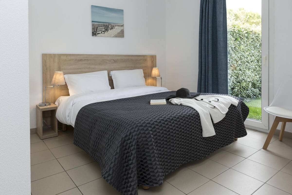 A neatly made bed with a dark patterned bedspread, two white pillows, and a wooden headboard. There are two bedside lamps on either side of the bed. A small table with a lamp is visible to the left of the bed.
