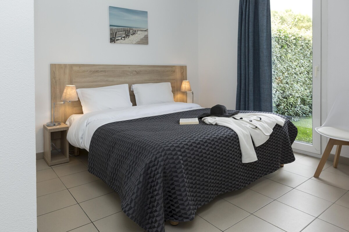 A neatly made bed with a geometric patterned bedspread, two bedside lamps, a small bedside table, and a window with a view of greenery.
