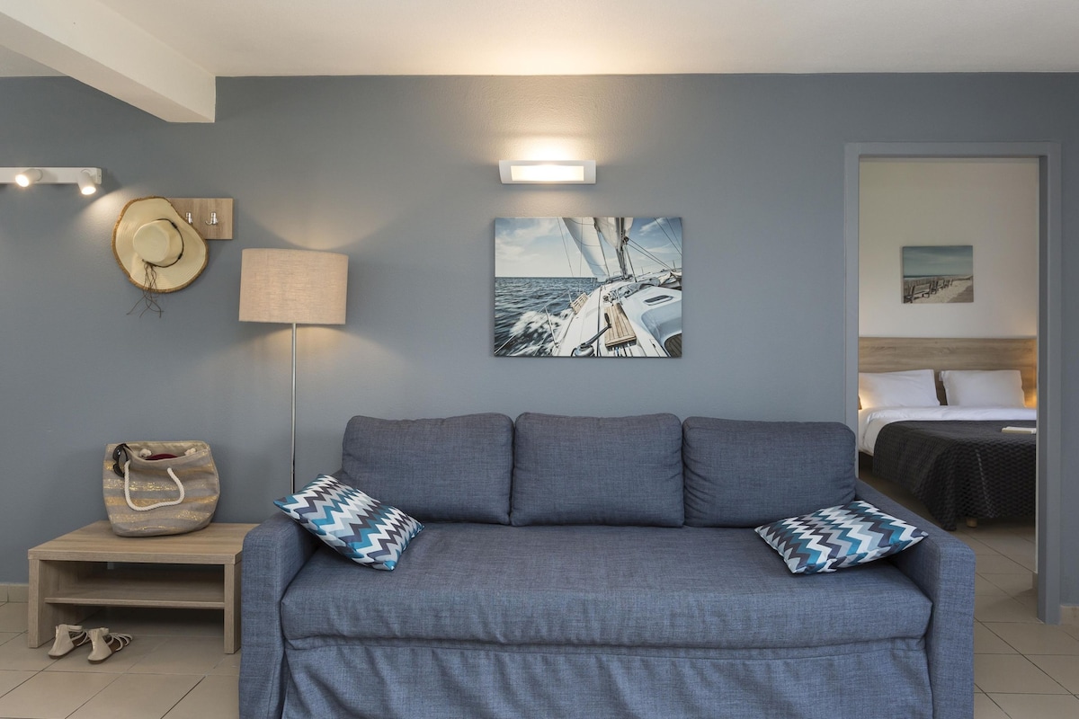 A room with a grey sofa, two patterned pillows, a wall-mounted lamp, a hat, a handbag, and a framed picture of a sailboat.