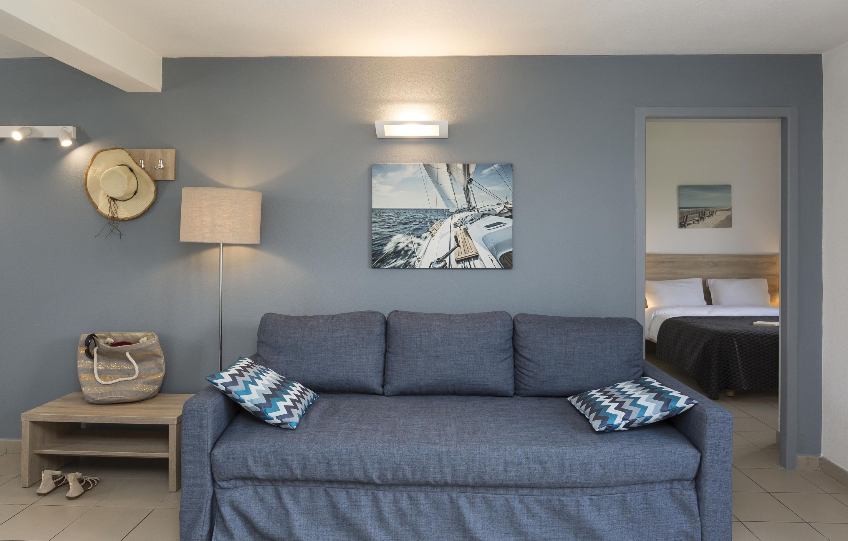 A room with a grey sofa, two patterned pillows, a wall-mounted lamp, a hat, a handbag, and a framed picture of a sailboat.