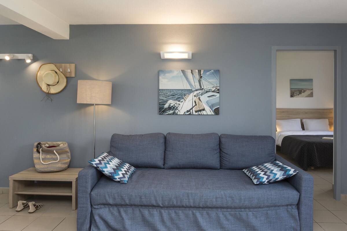 A room with a grey sofa, two patterned pillows, a wall-mounted lamp, a hat, a handbag, and a framed picture of a sailboat.