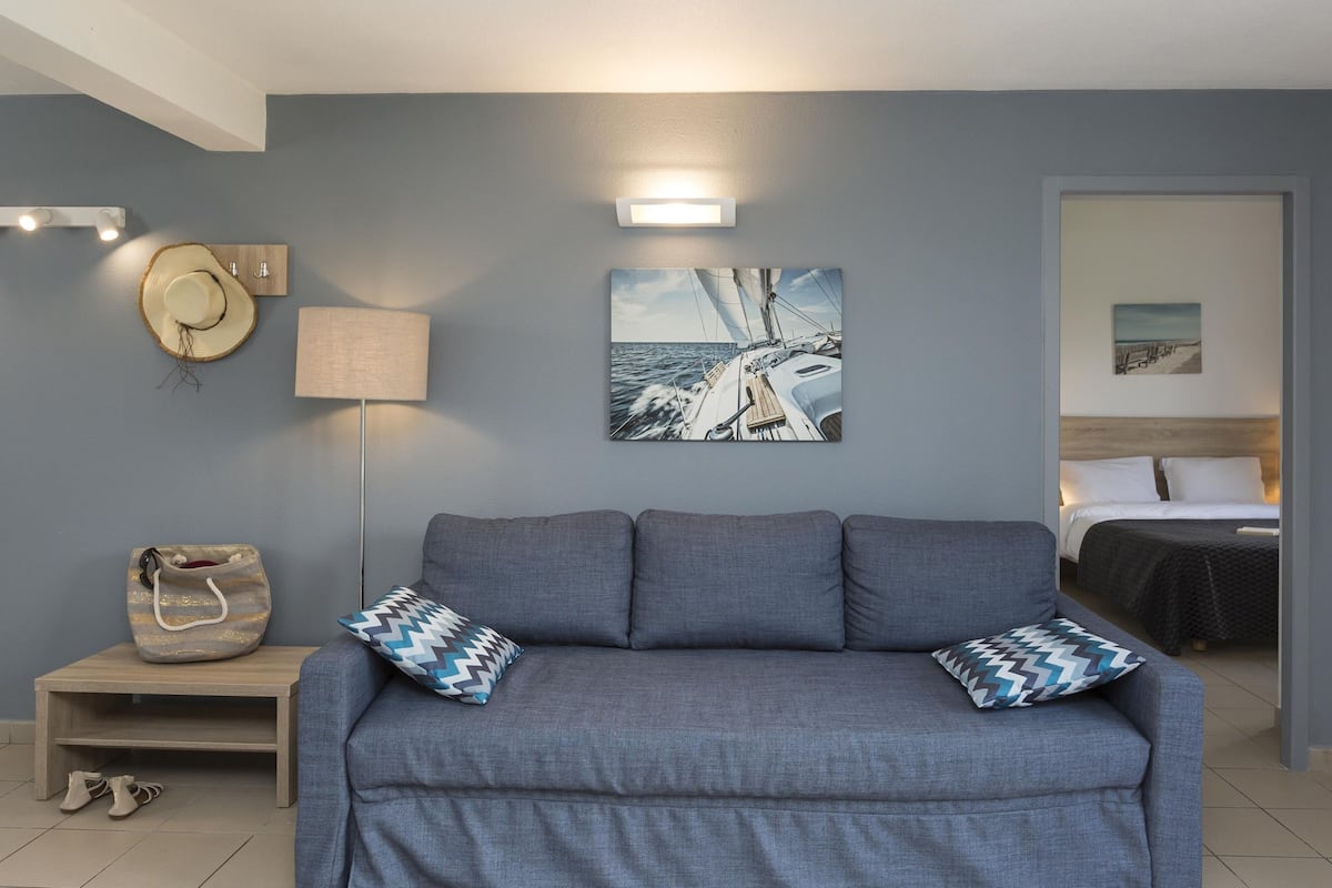 Una habitación con un sofá gris, dos almohadones estampados, una lámpara de pared, un sombrero, un bolso y un cuadro enmarcado de un velero.