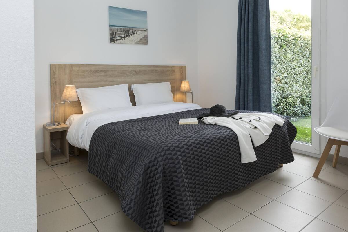 A neatly made bed with a geometric patterned bedspread, two bedside lamps, a small bedside table, and a window with a view of greenery.