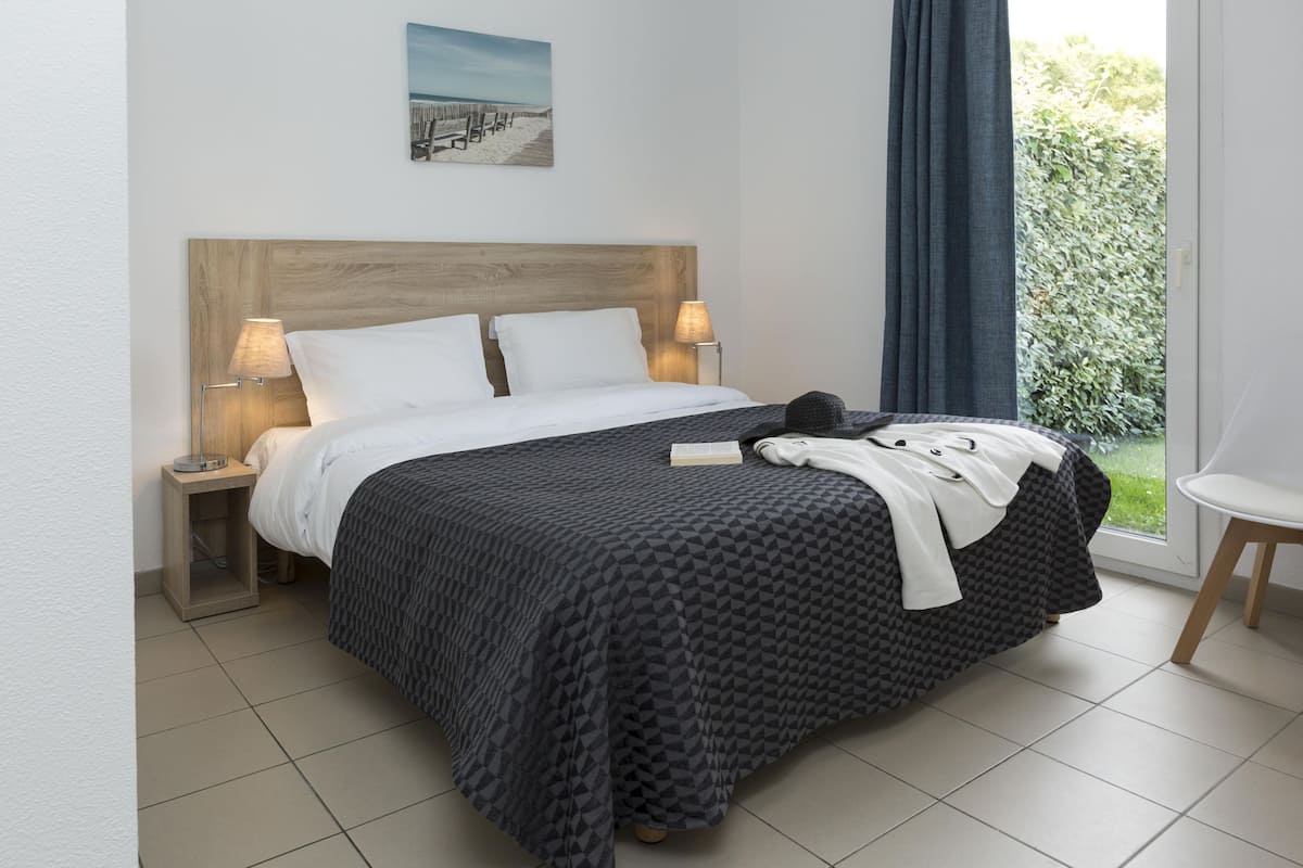 Una cama bien hecha con una colcha de estampado geométrico, dos lámparas de noche, una mesita de noche pequeña y una ventana con vistas a la vegetación.