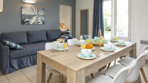 A dining area with a wooden table set for breakfast, a grey sofa, and a painting of a sailboat on the wall. - Résidence Odalys Le Village des Amareyeurs (Le Chateau-d'Oleron)