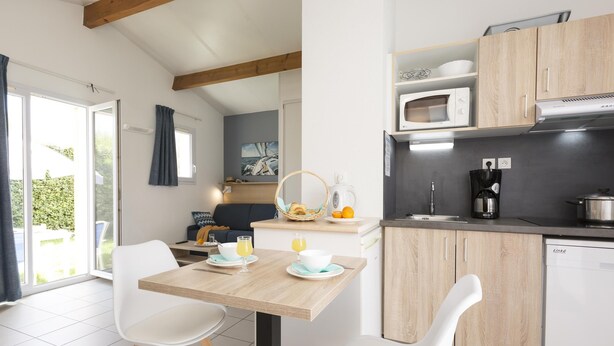 A modern kitchen with wooden cabinets, a dining table with chairs, and a breakfast setup with orange juice. - Résidence Odalys Le Village des Amareyeurs (Le Chateau-d'Oleron)
