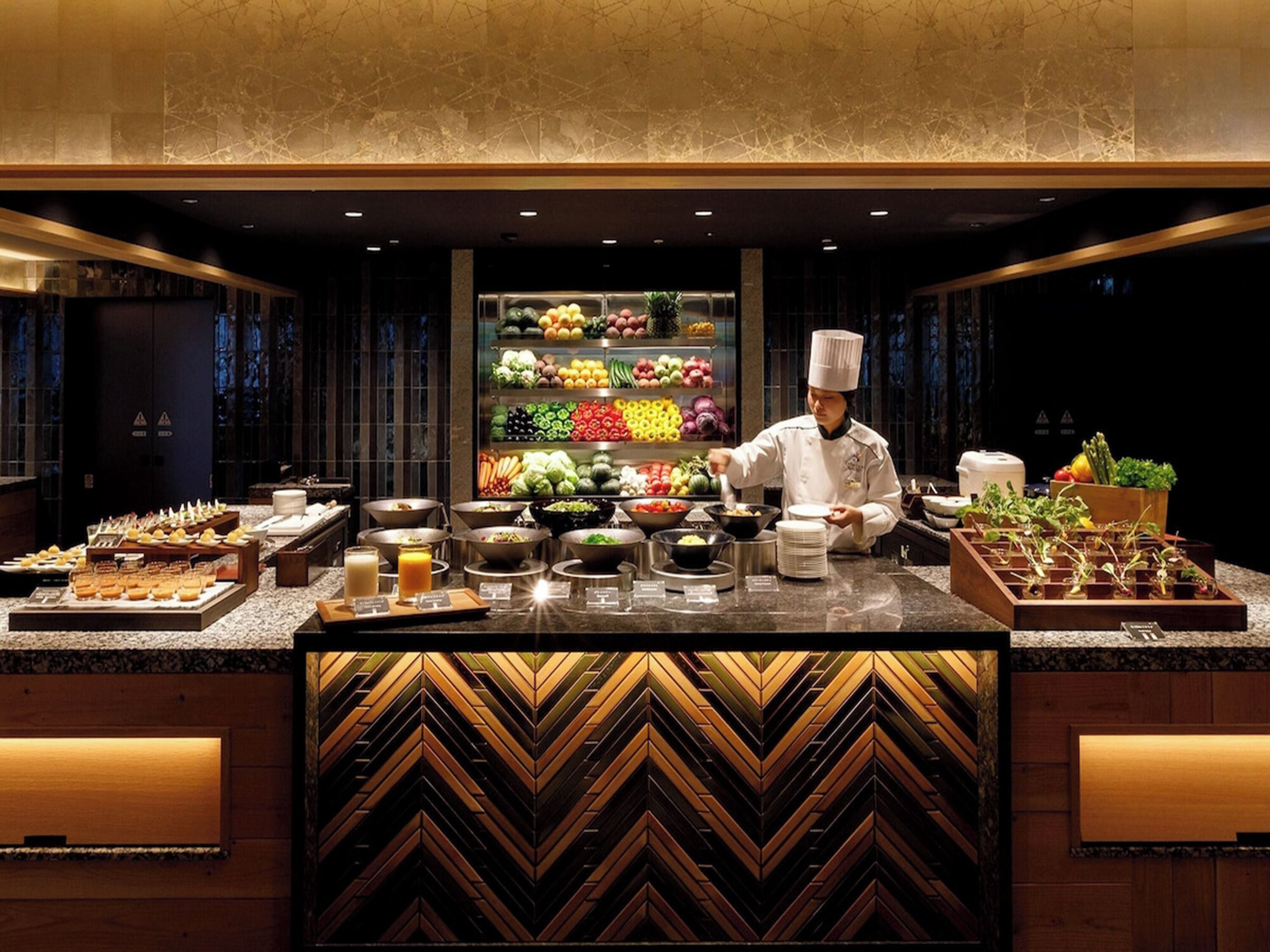 A chef preparing food in a modern kitchen with a variety of fruits and vegetables displayed in the background.