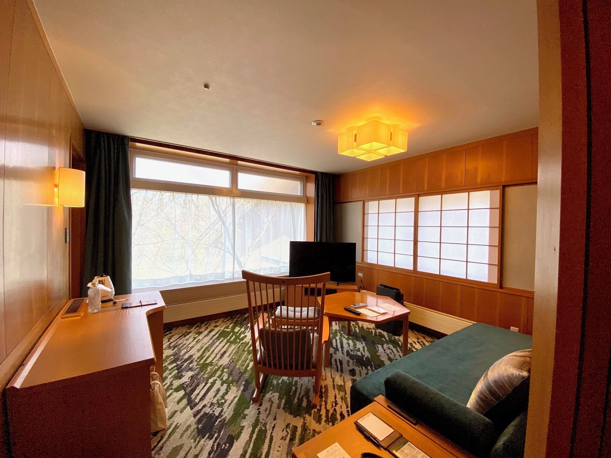 A hotel room with a bed, a desk with a lamp, a chair, a television, and a large window with curtains.
