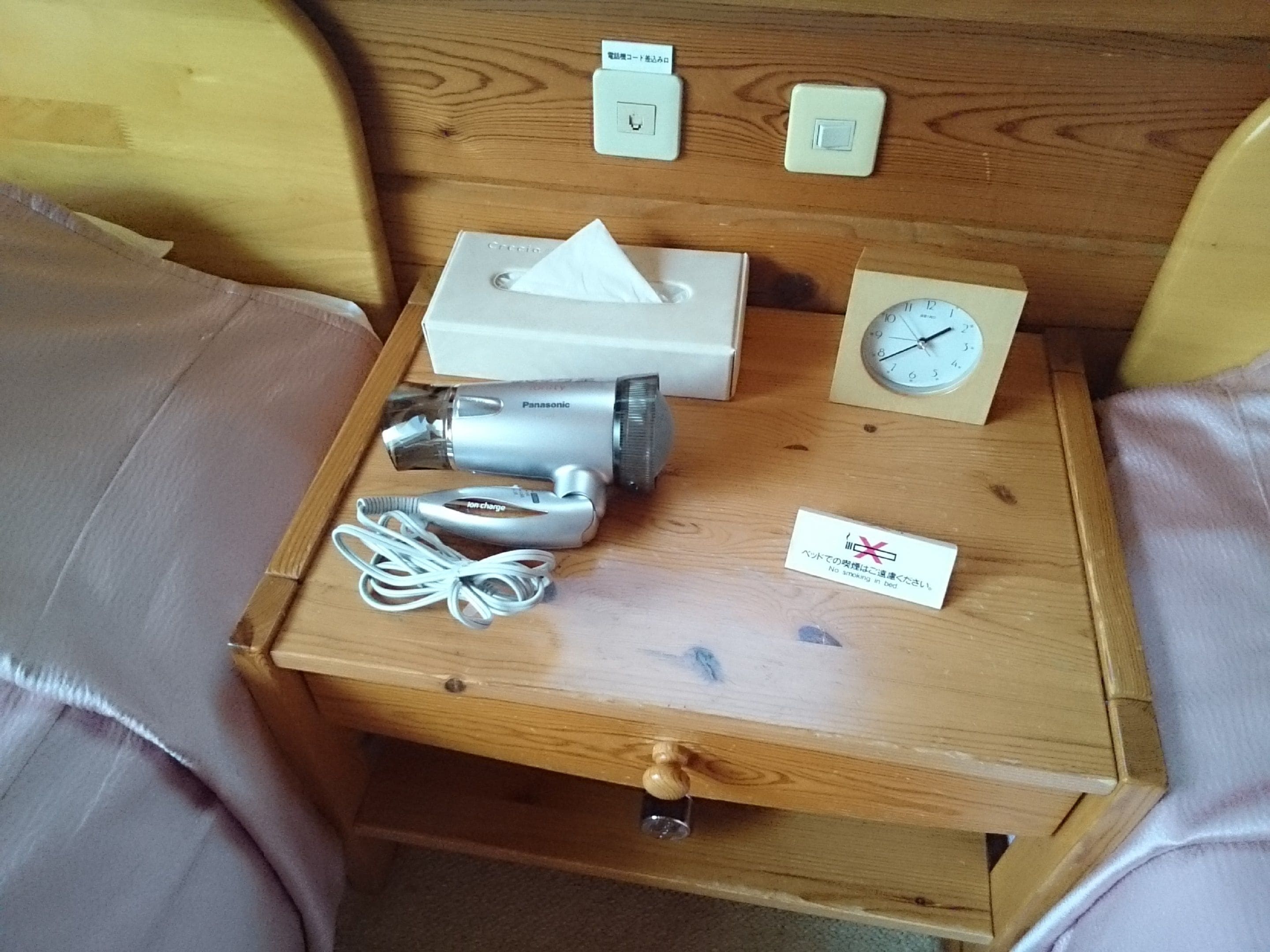 A wooden bedside table with a Panasonic hairdryer, a tissue box, a clock, and a no-smoking sign.