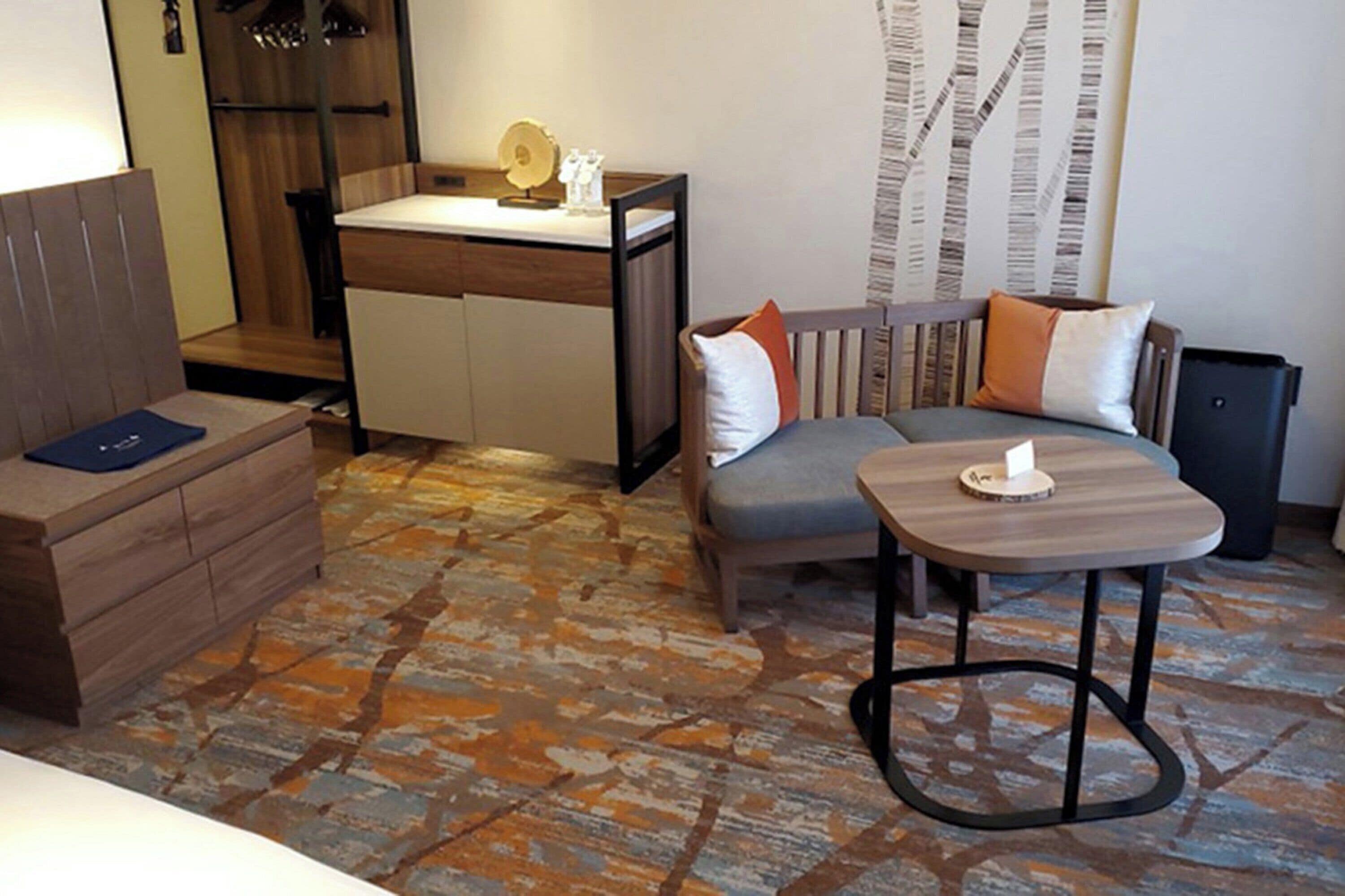 A modern hotel room with a wooden bench, a small table, a sofa with cushions, and a patterned floor.
