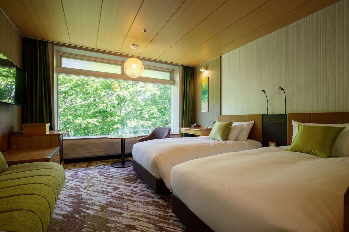 Une chambre d’hôtel avec deux lits, un bureau, une chaise, une grande fenêtre donnant sur des arbres et un luminaire au plafond.