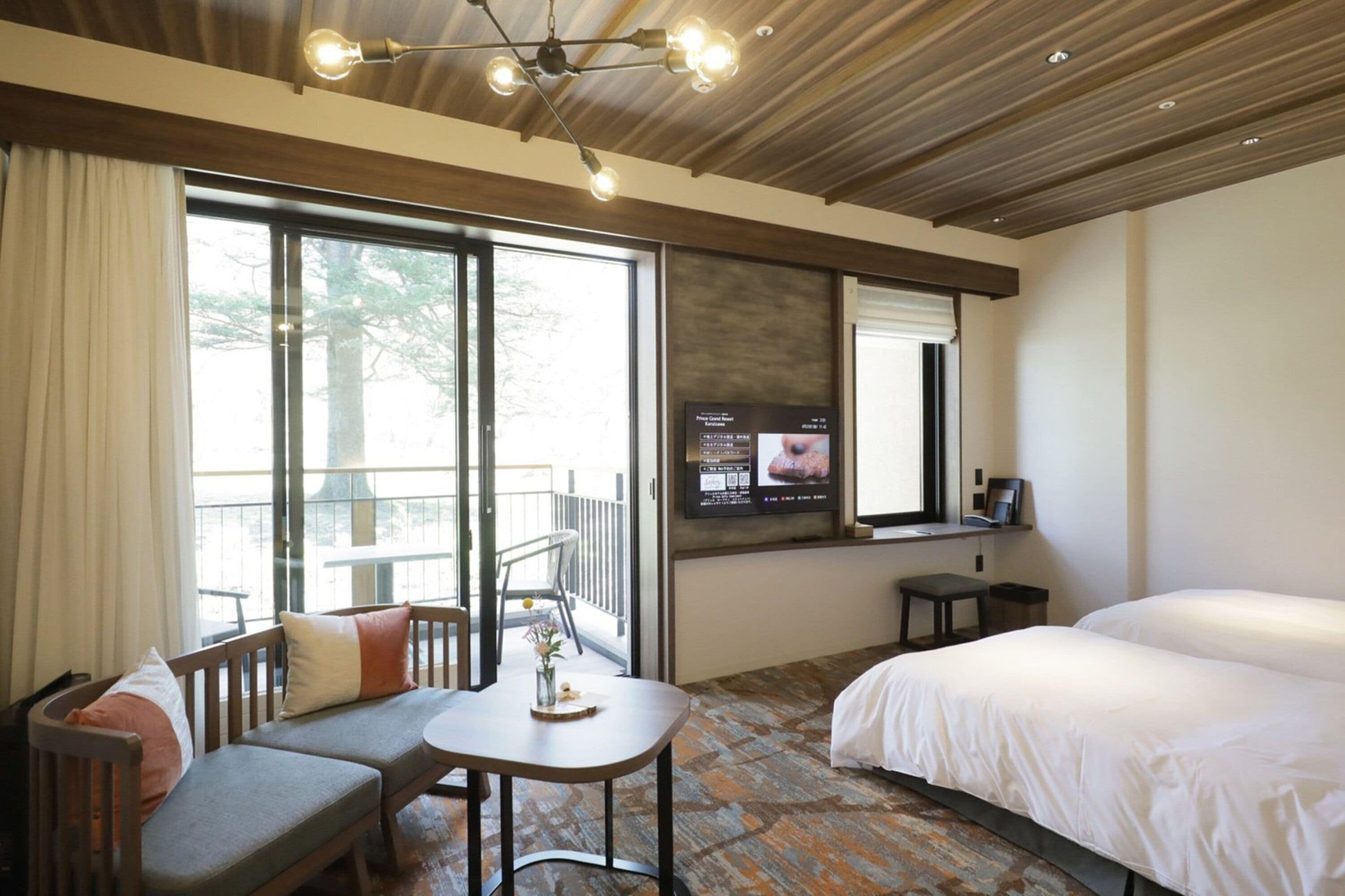 A hotel room with a bed, a sofa, a small table, a TV, and a balcony with a view of trees.