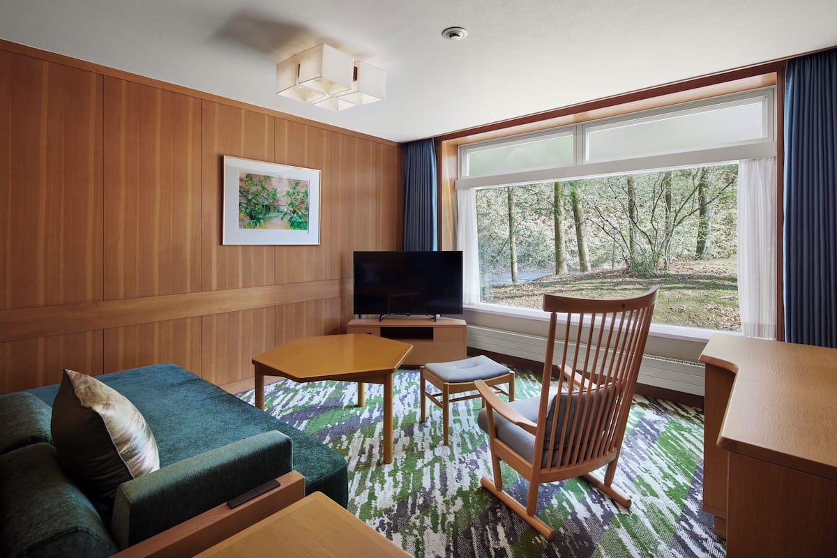 Une chambre d’hôtel avec un mur lambrissé en bois, un téléviseur à écran plat, un canapé vert, une chaise en bois, une petite table et une grande fenêtre donnant sur des arbres.