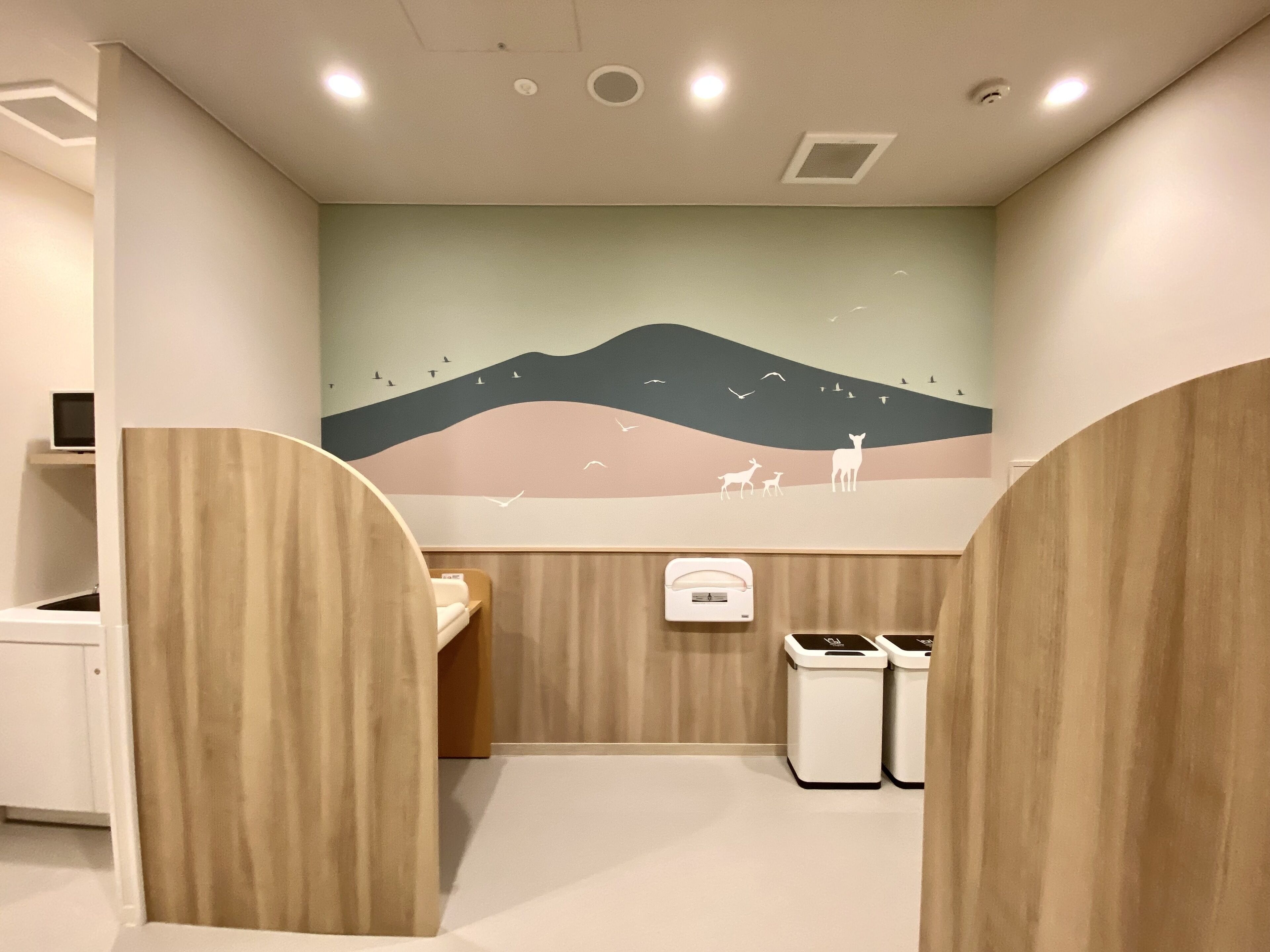 A children's bathroom with a mural of a landscape, wooden partitions, and a trash bin.