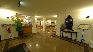 Um saguão de hotel com piso lustroso, balcão de recepção, planta em vaso, relógio e extintor de incêndio.
