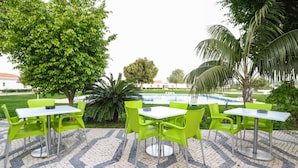 Uma área de varanda ao ar livre com cadeiras verdes e mesas brancas, cercada por árvores e uma piscina.