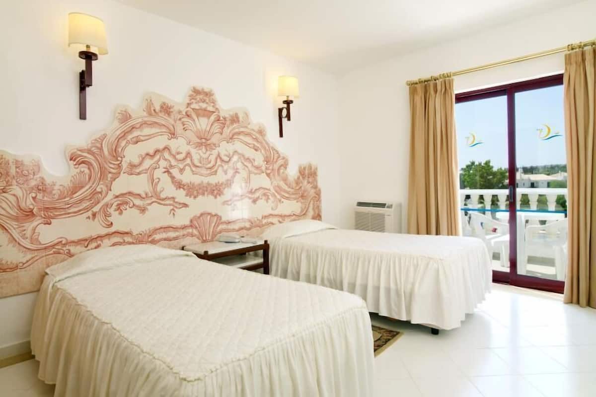 A hotel room with two beds, a decorative headboard, wall-mounted lamps, an air conditioning unit, and a balcony with curtains.