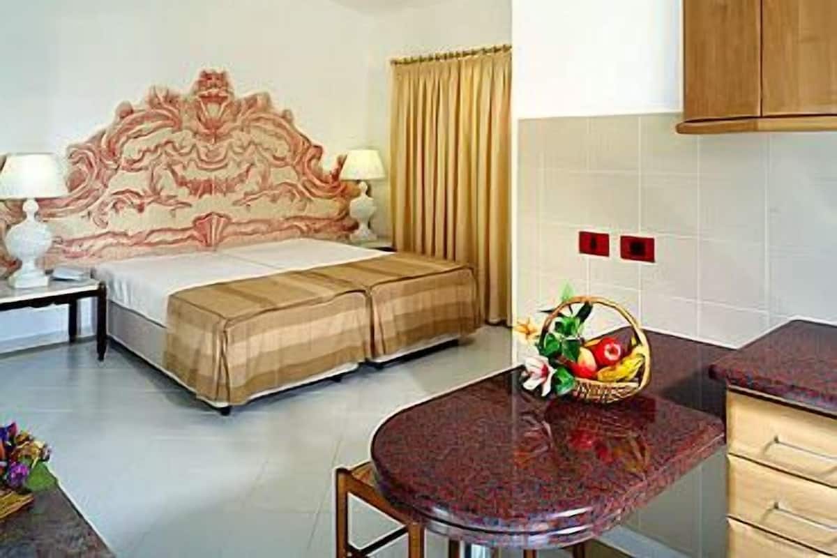 A hotel room with a bed, a bedside table with a lamp, a small table with a fruit basket, and a kitchen area with cabinets.