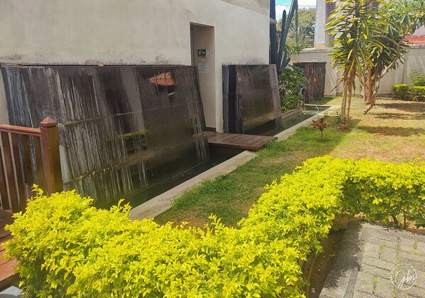 A courtyard with a small water feature, neatly trimmed bushes, and a building with a dark facade. - Grand Bay Suites (Grand-Baie)