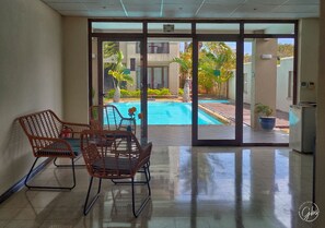 A spacious indoor area with a large sliding glass door leading to an outdoor swimming pool. There are wicker chairs and a small table in the room. - Grand Bay Suites (Grand-Baie)