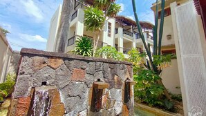 A stone wall with a water feature, surrounded by tropical plants and cacti, with a multi-story building in the background. - Grand Bay Suites (Grand-Baie)