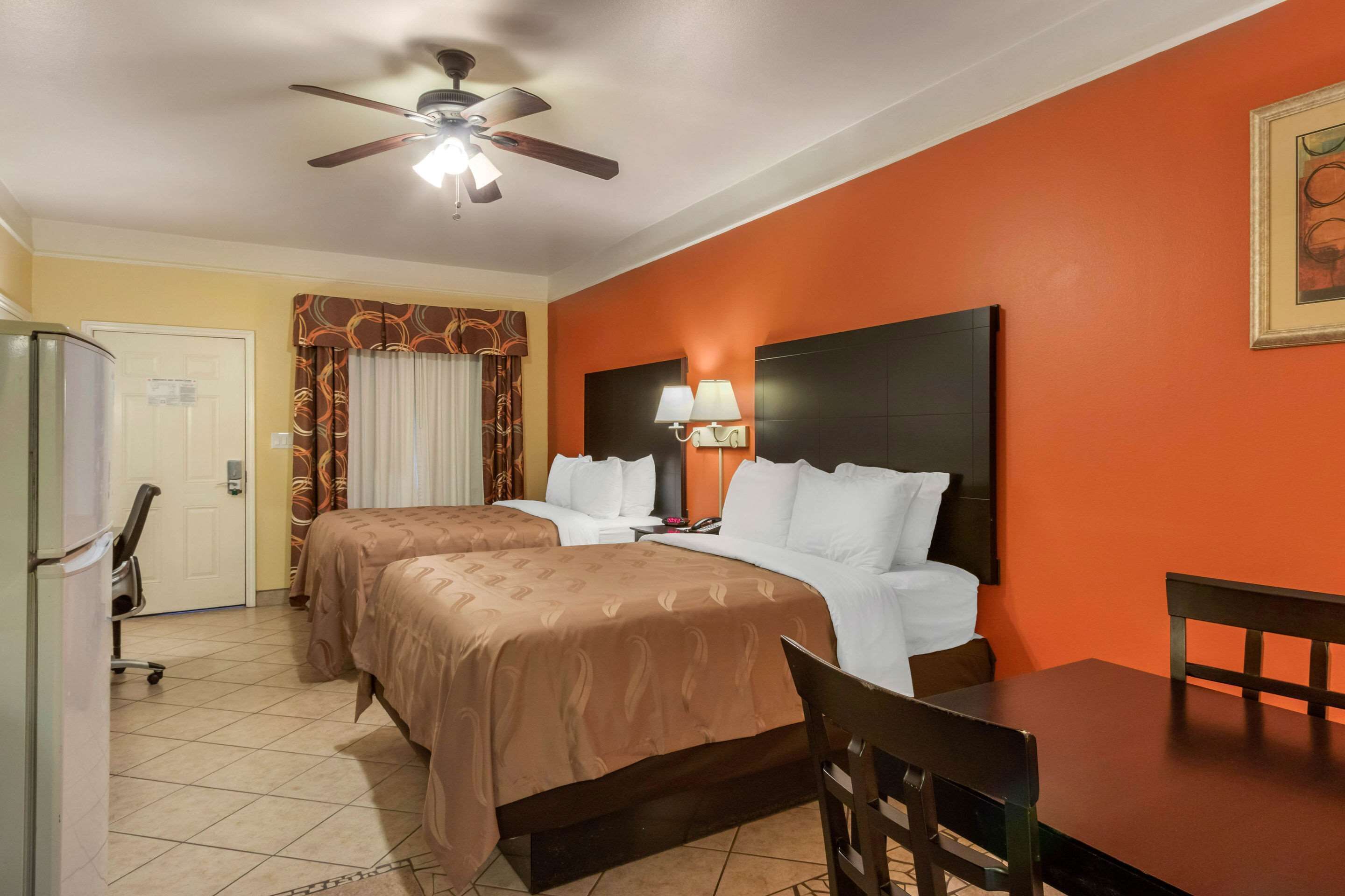 A hotel room with two beds, a desk, a chair, a ceiling fan, and a wall-mounted lamp.