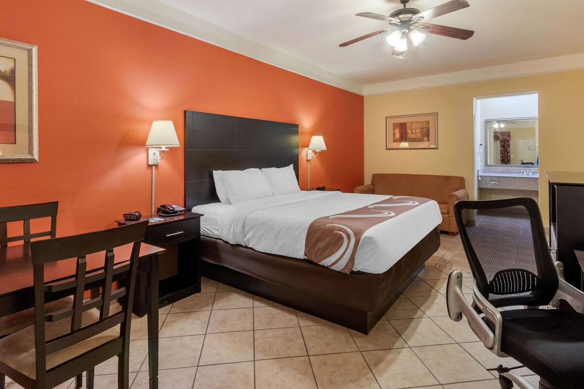 A hotel room with a large bed, a desk with a chair, a desk lamp, and a ceiling fan.