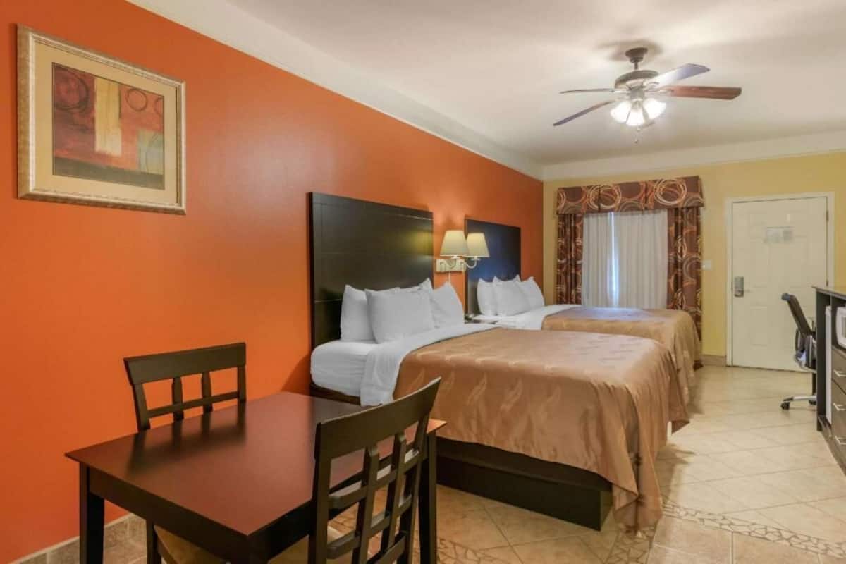 A hotel room with two beds, a desk, chairs, a ceiling fan, and a door.