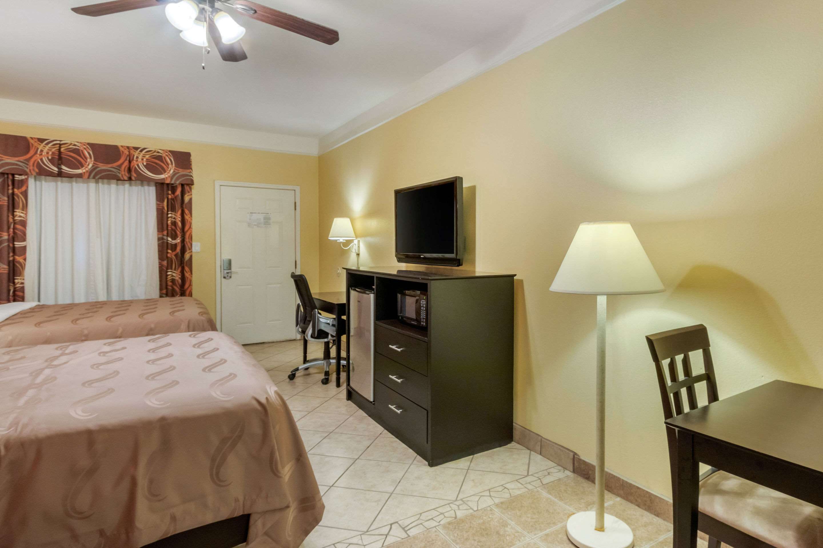 A hotel room with two beds, a desk, a television, a lamp, and a ceiling fan.