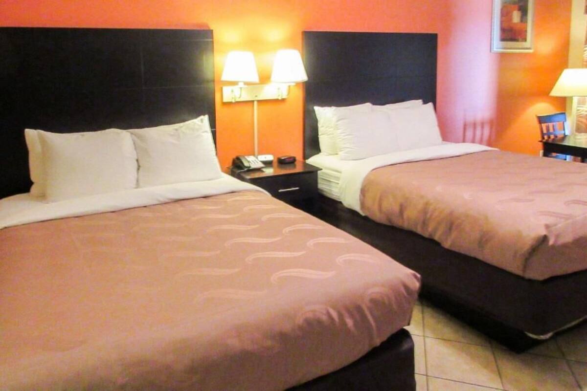 A hotel room with two beds, a nightstand with a lamp, and a wall-mounted lamp.