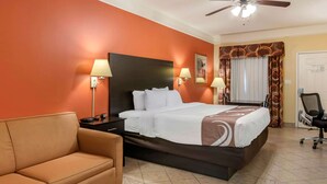 A hotel room with a bed, a nightstand, a lamp, a sofa, a ceiling fan, and a door. - Quality Inn & Suites at The Outlets Mercedes/Weslaco (Mercedes)