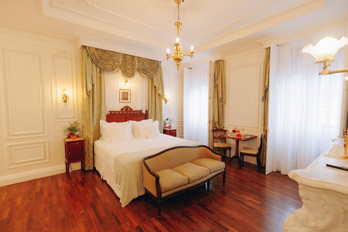 Een ruime slaapkamer met een groot bed, een houten bankje, een klein tafeltje en een decoratieve spiegel.