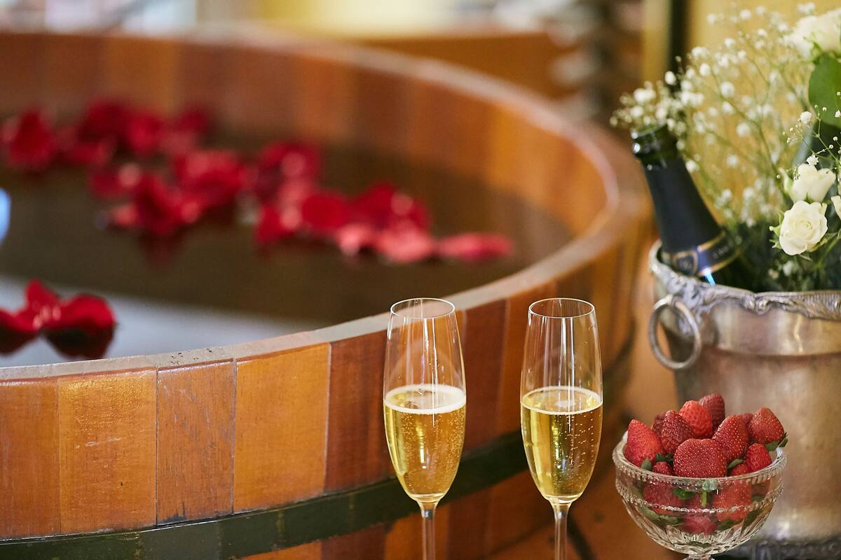 Twee champagneglazen op een reflecterend oppervlak, een schaal met aardbeien en een zilveren karaf met bloemen.