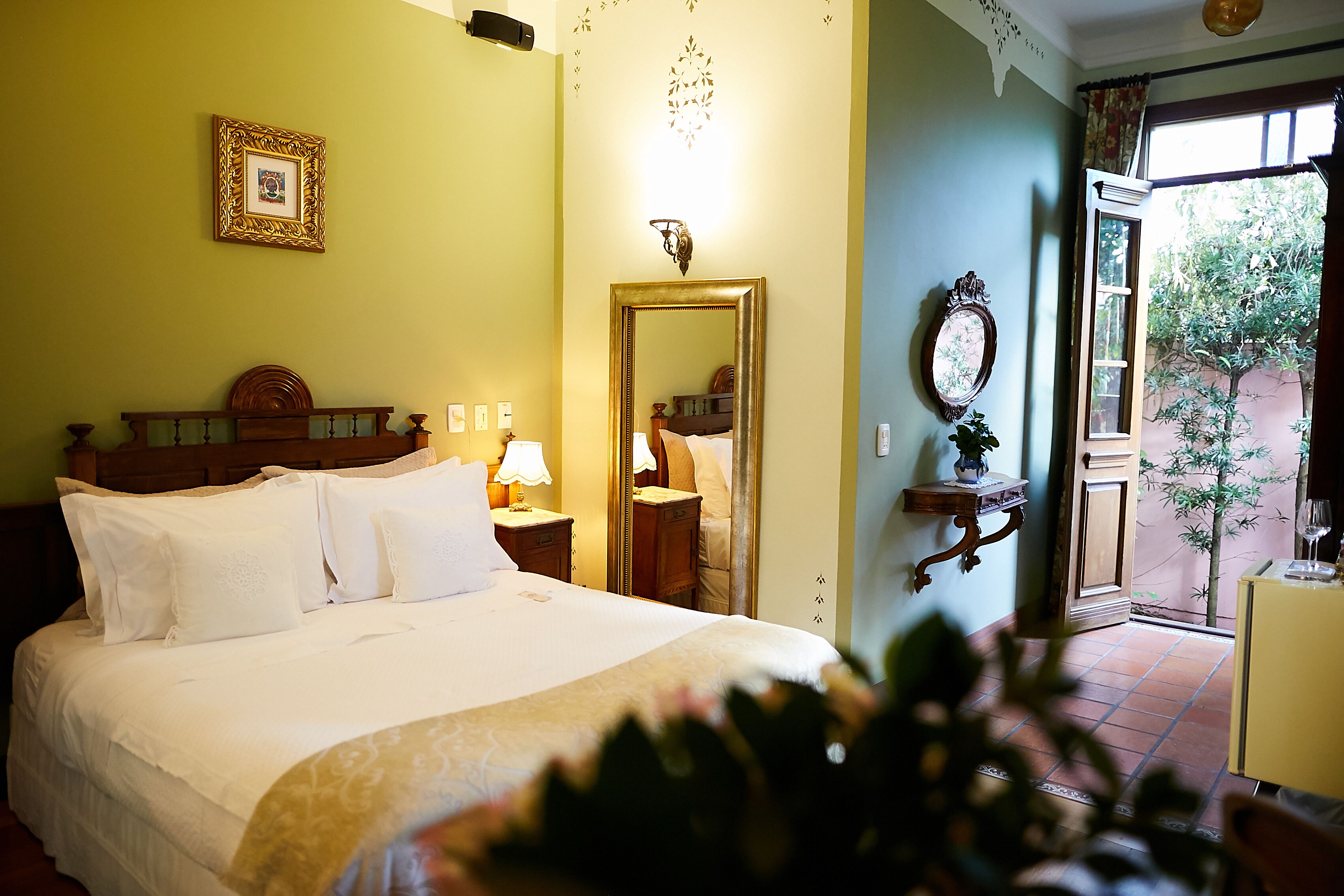 A hotel room with a large bed, a bedside table with a lamp, a mirror, and a window with a view of greenery outside.