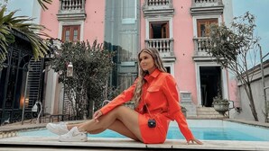 A woman sitting by a pool in front of a pink building with balconies and a tall central section.
