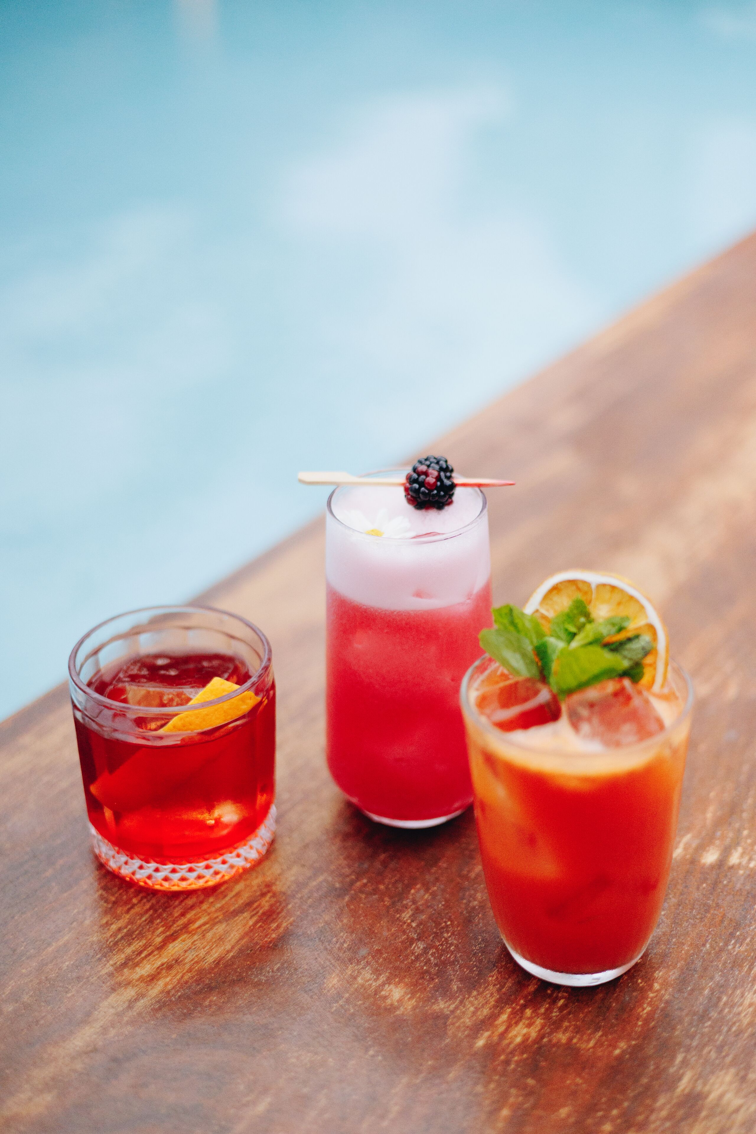Trois verres de boissons rouges, décorés de fruits ou d’autres éléments, sont posés sur une surface en bois, près d’une piscine.