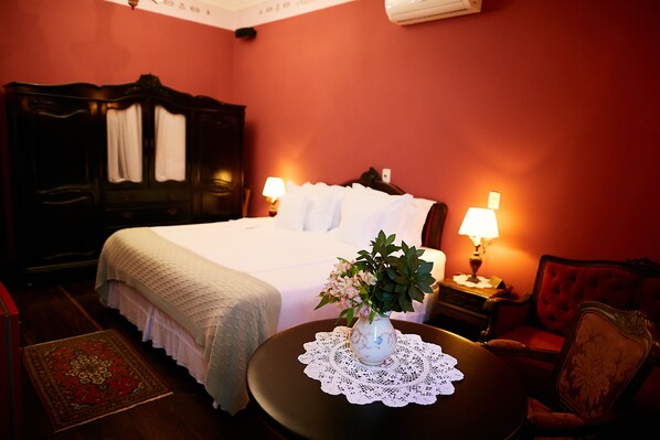 Habitación de hotel con una cama grande, dos lámparas de noche, un jarrón con flores, una silla roja y una mesita redonda con un mantel de encaje.